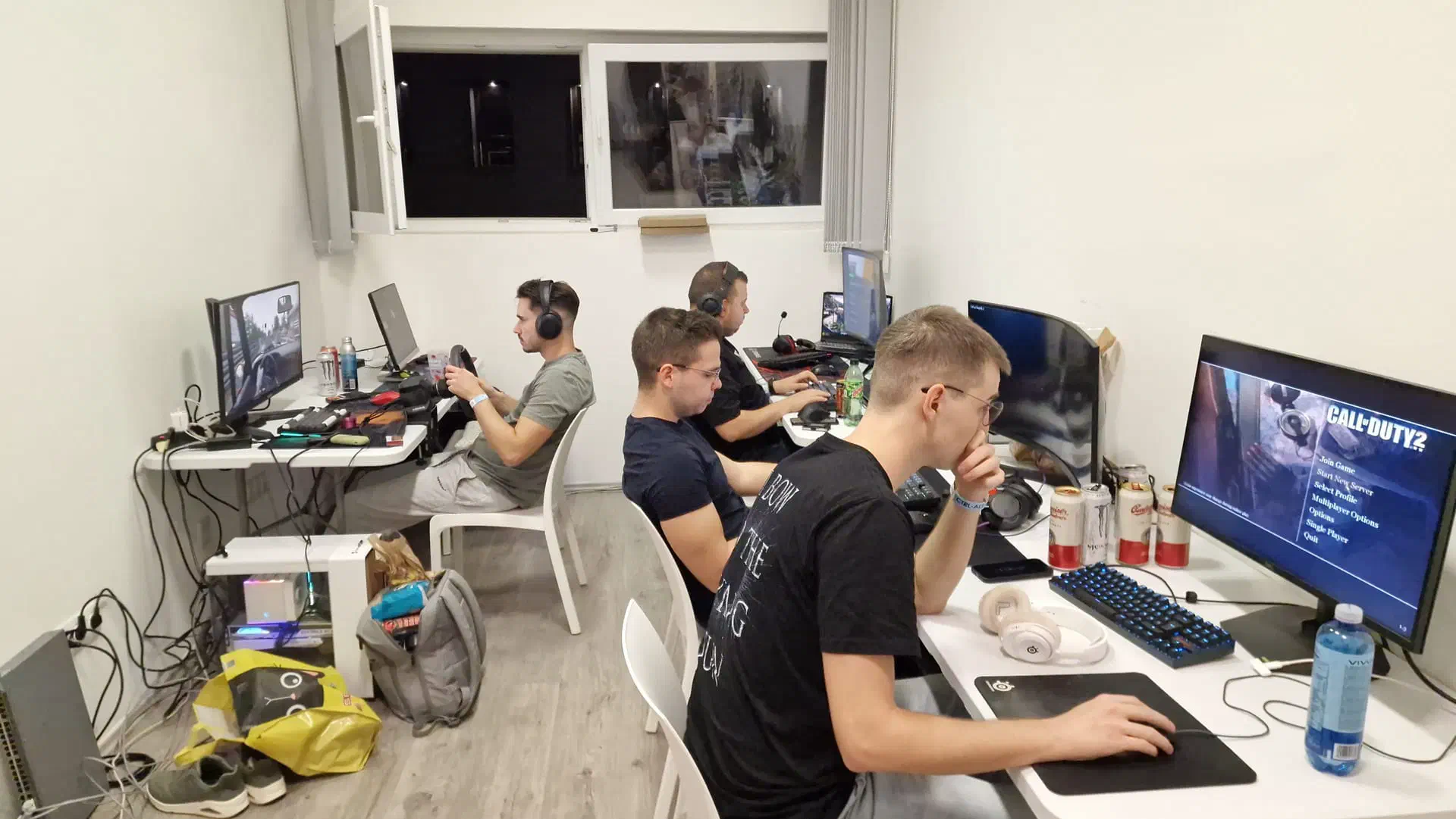 Este a keskeny teremben: elöl egy szemüveges játékos a Call of Duty 2 főmenüjét nézi a monitoron, mögötte többen játszanak a fal menti asztaloknál